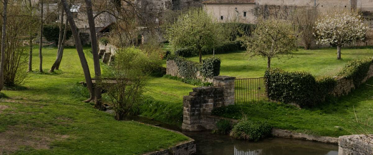 A historic stone castle with ruins, a small stream, and a well-maintained garden with trees and a hedge.