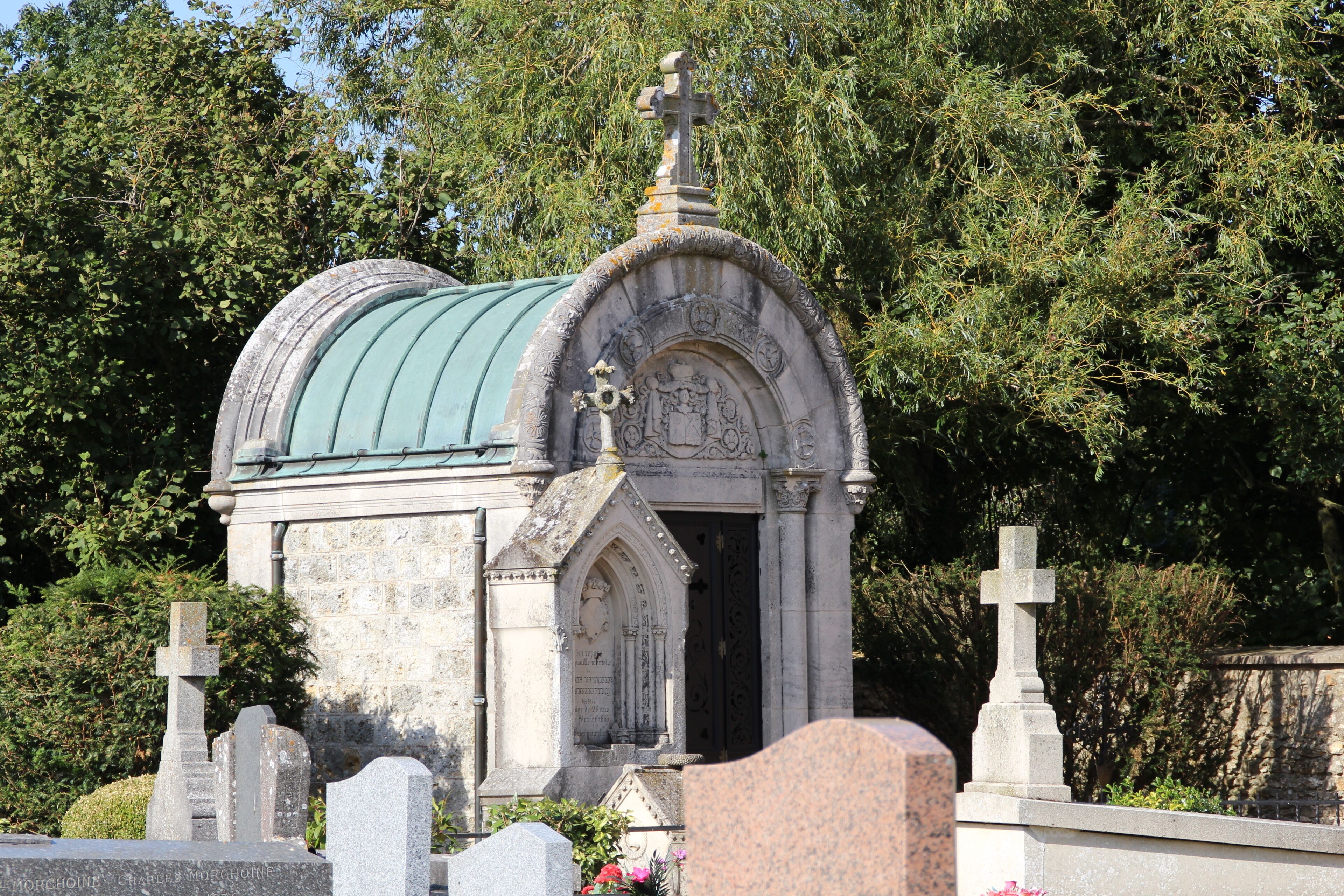A small stone chapel with a green roof, surrounded by trees and other gravestones.