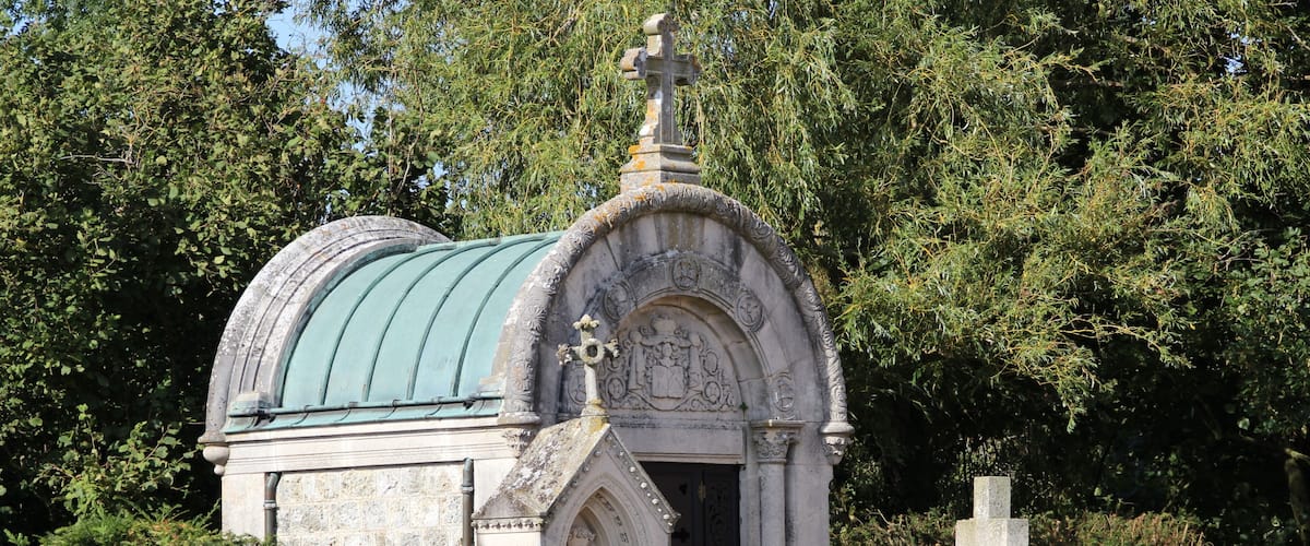 A small stone chapel with a green roof, surrounded by trees and other gravestones.