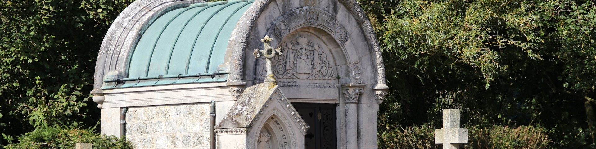 A small stone chapel with a green roof, surrounded by trees and other gravestones.