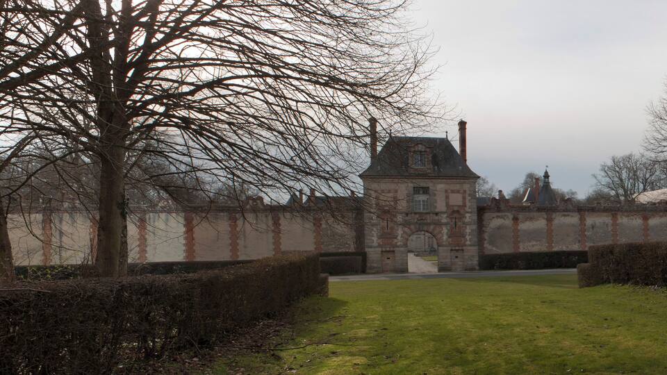 A historic building with a tower, surrounded by hedges and leafless trees.