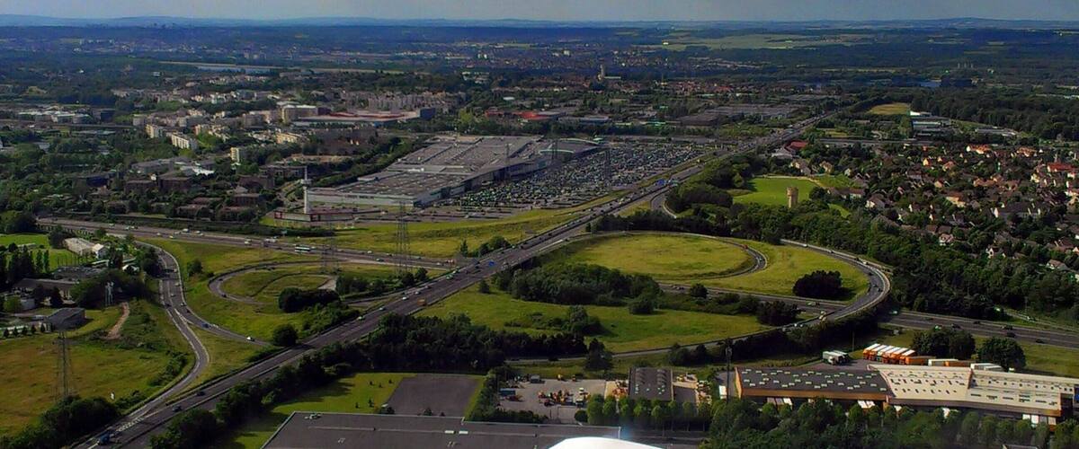 An aerial view of a city with a large industrial area, multiple highways, and surrounding green spaces.