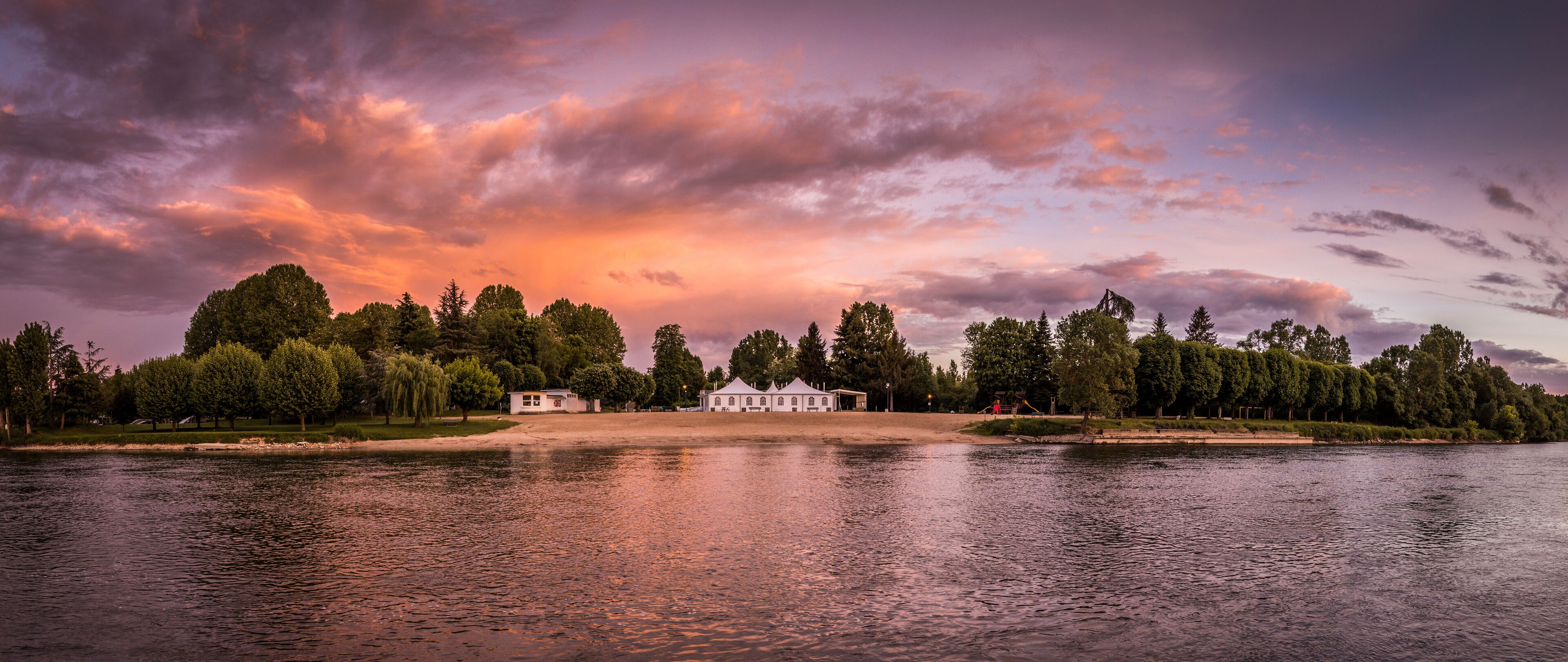 A lakeside view with a white building and trees under a colorful sunset sky.