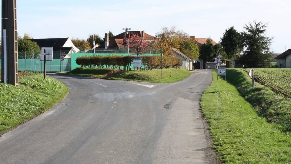 A rural road with a sharp bend, surrounded by greenery and houses.