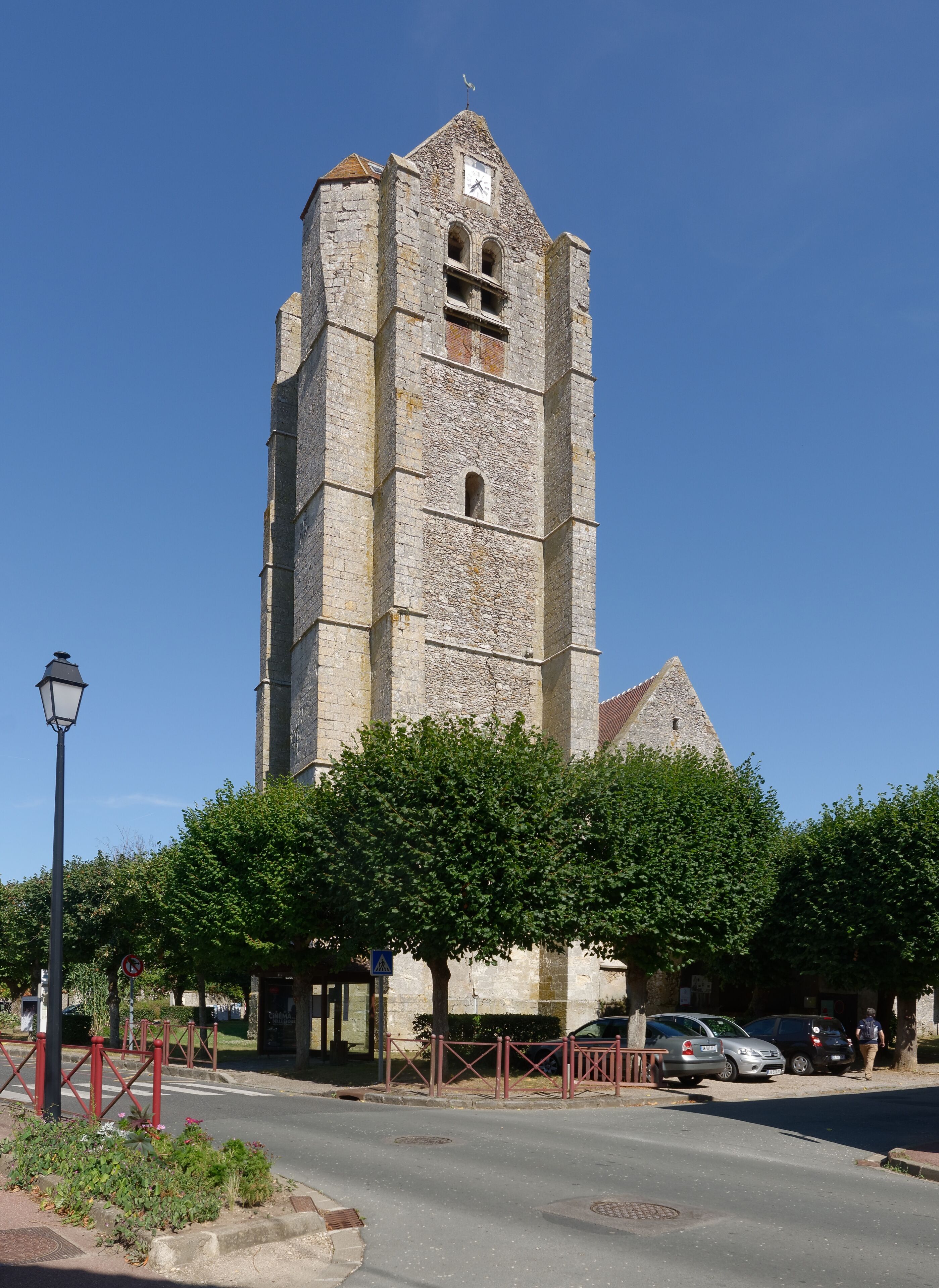 A tall stone church with a clock tower, surrounded by trees and parked cars.