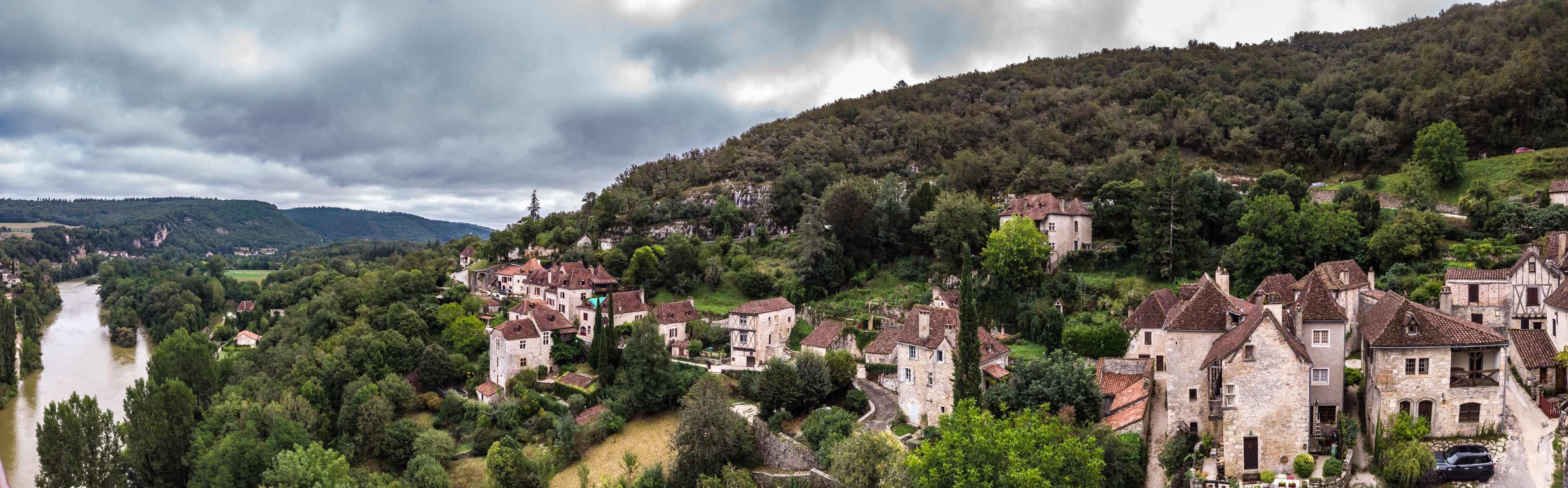 A picturesque village with stone houses nestled by a river, surrounded by lush greenery and hills.