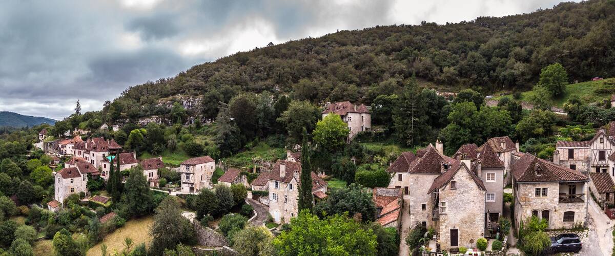 A picturesque village with stone houses nestled by a river, surrounded by lush greenery and hills.