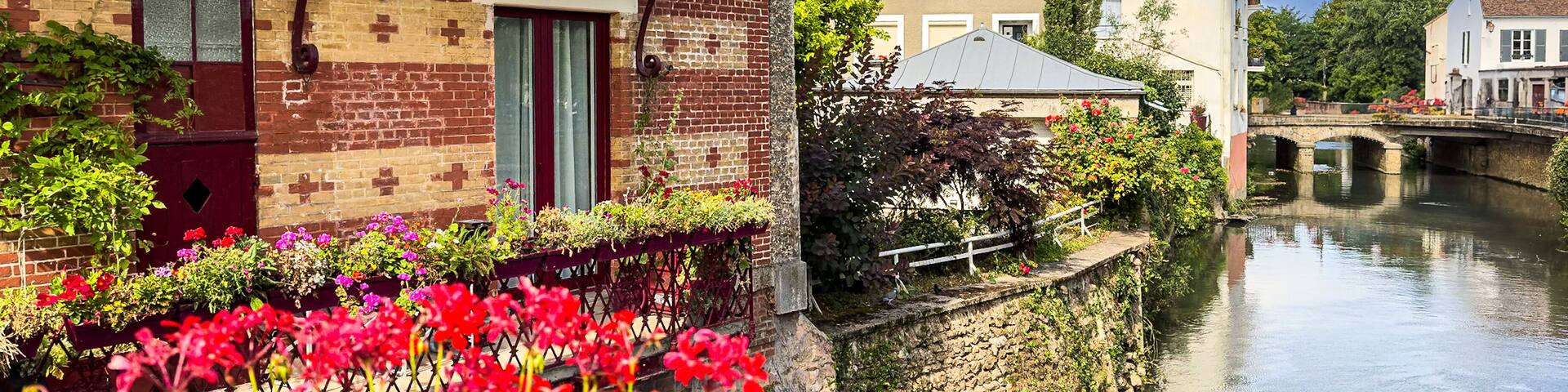 A canal with a bridge, a red balcony, and blooming flowers along the water's edge.