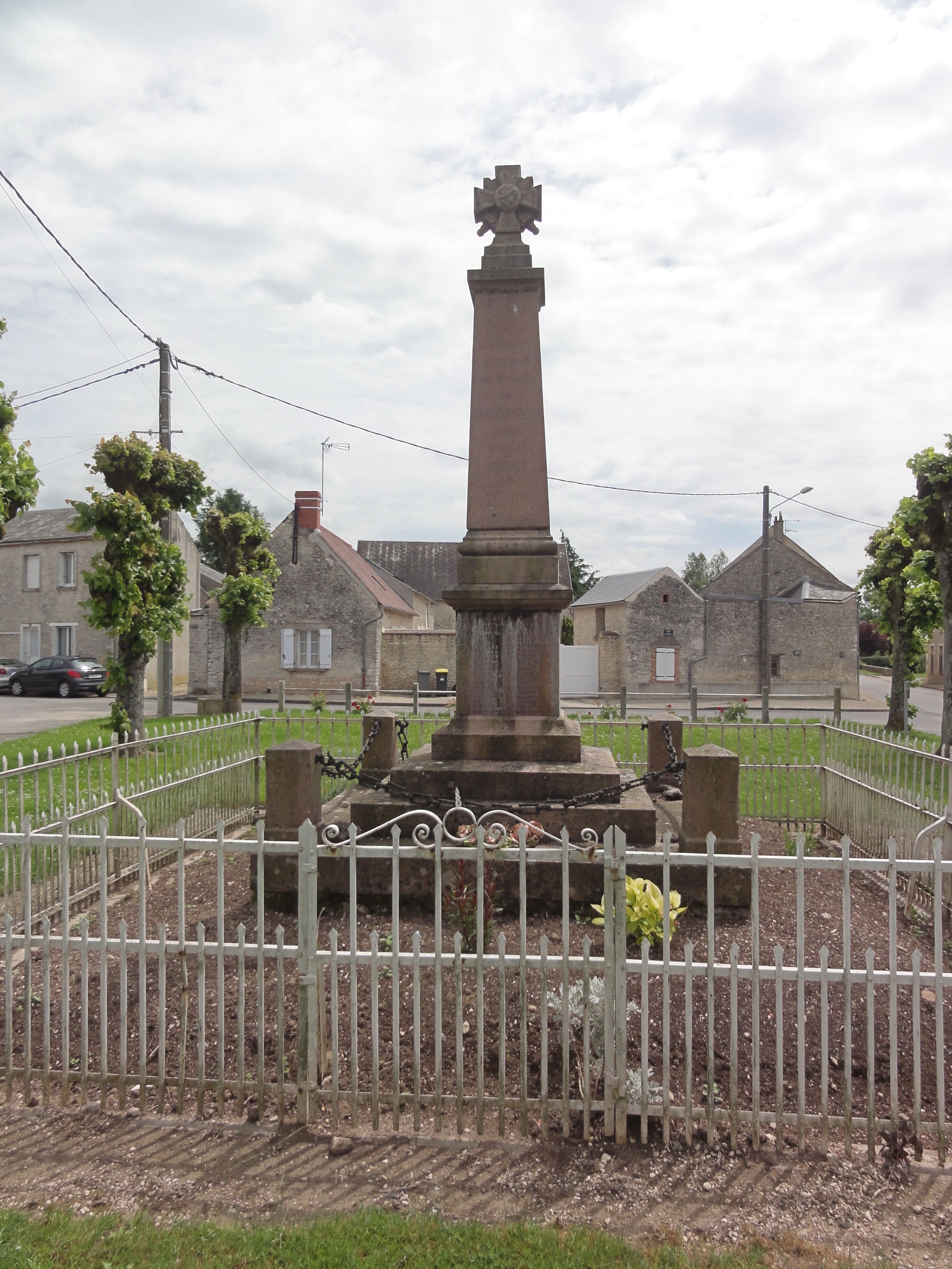 A tall stone monument with a cross on top, surrounded by a white fence and a grassy area.