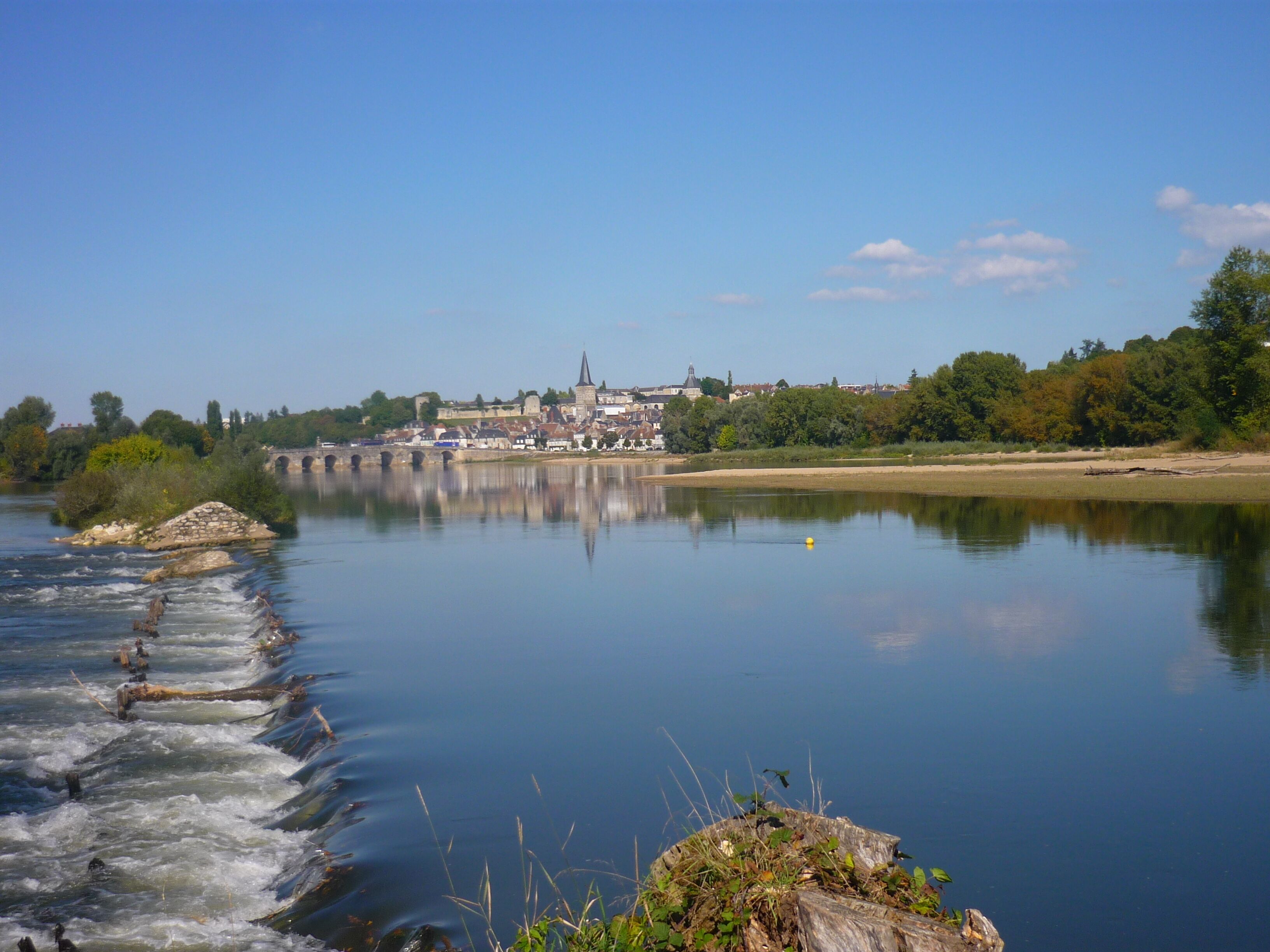A calm river with a bridge, a town in the background, and a forested area.