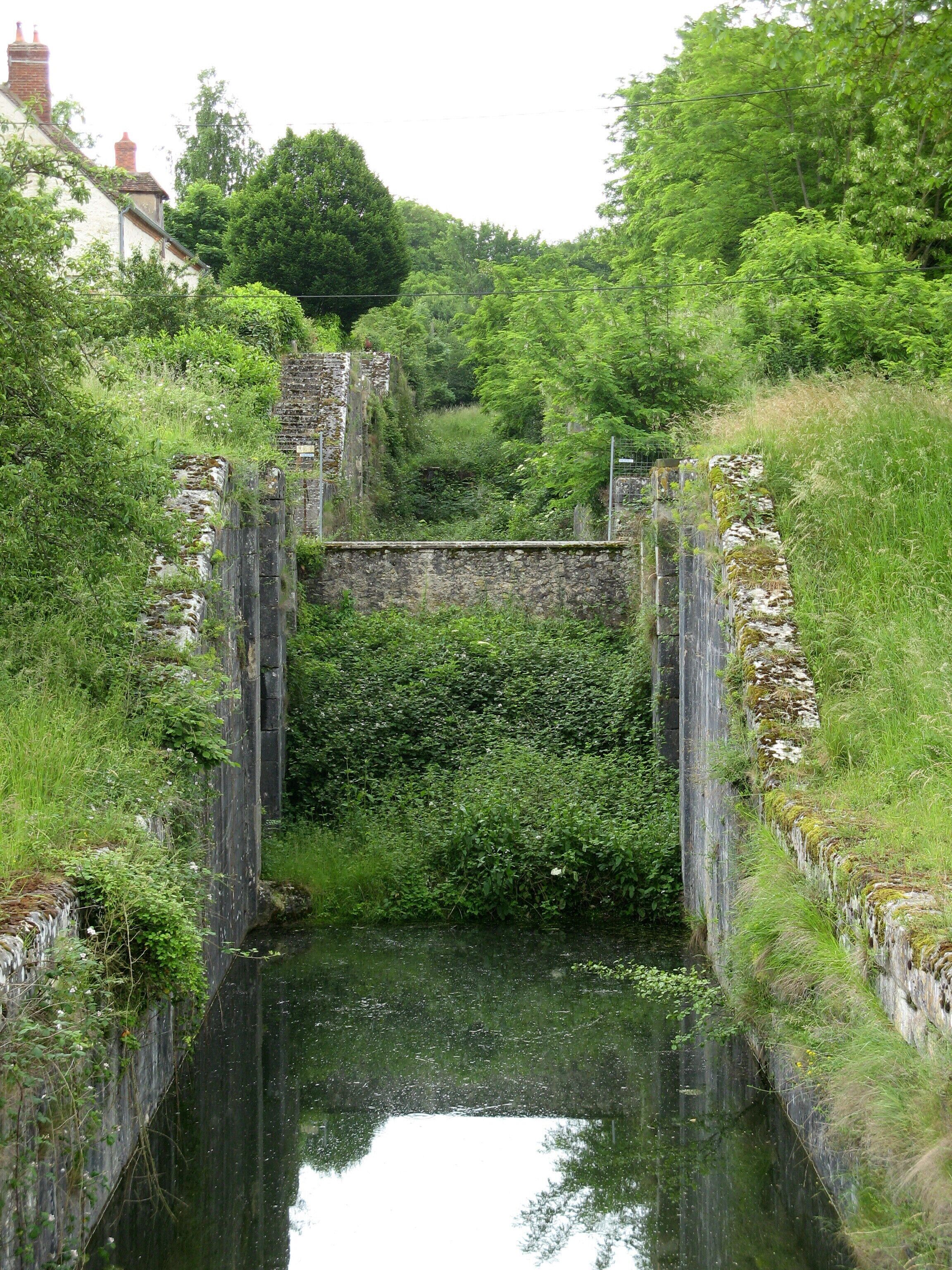 A narrow canal flanked by stone walls and lush greenery, with a small bridge crossing over the water.