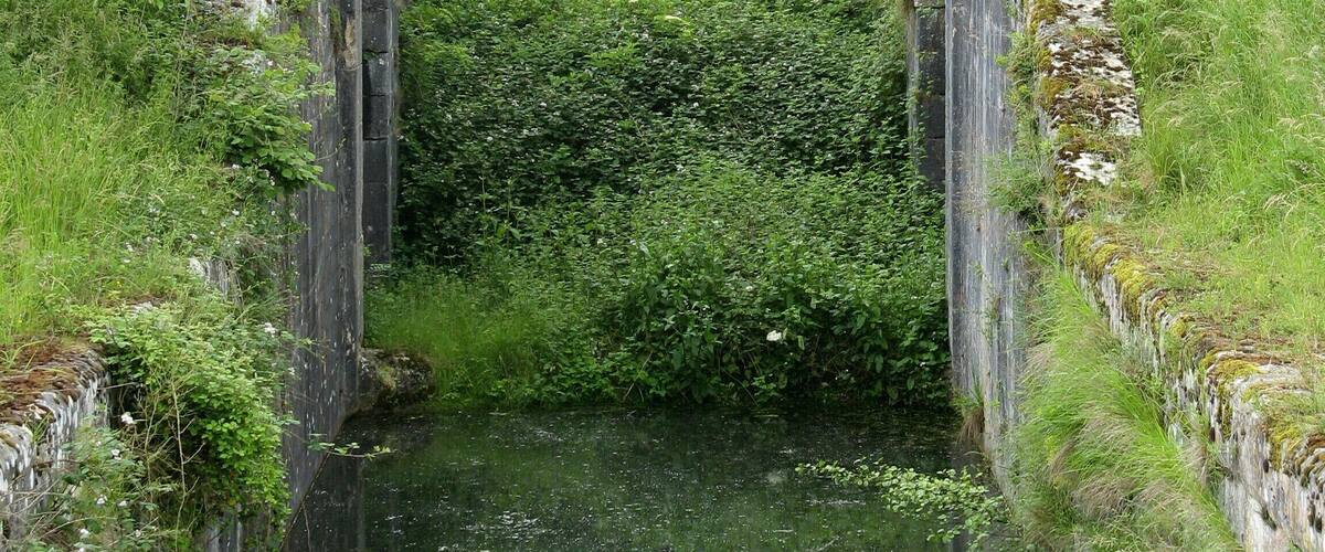 A narrow canal flanked by stone walls and lush greenery, with a small bridge crossing over the water.