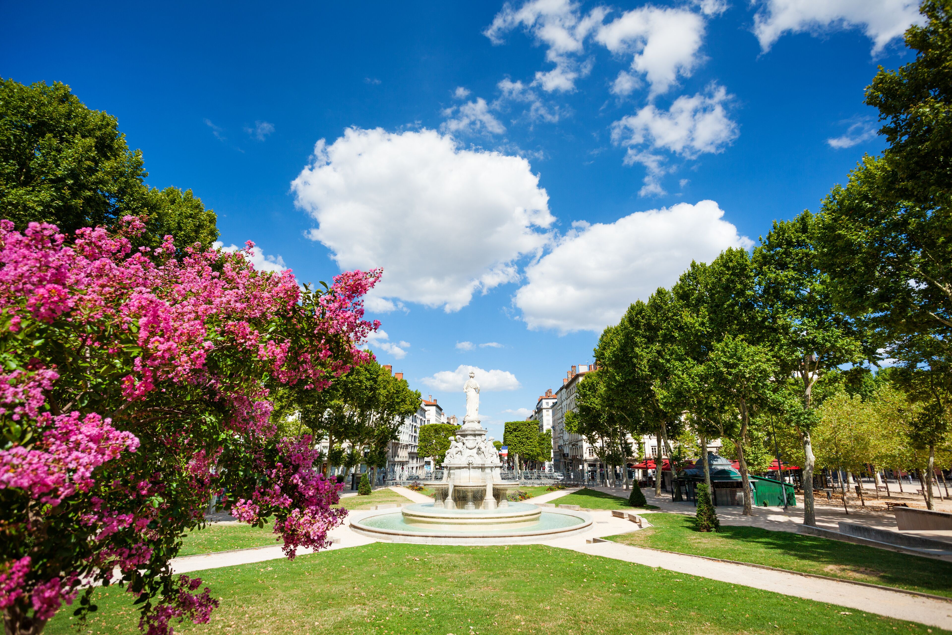 A park with a fountain, pink flowers, and a clear blue sky.