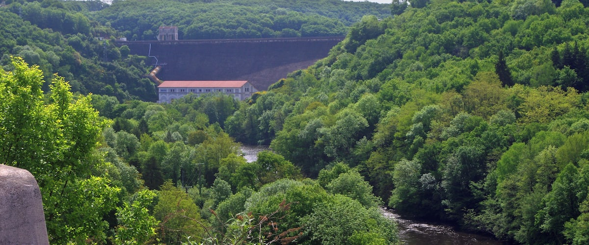 A lush green valley with a river, a dam, and a building on a hill.