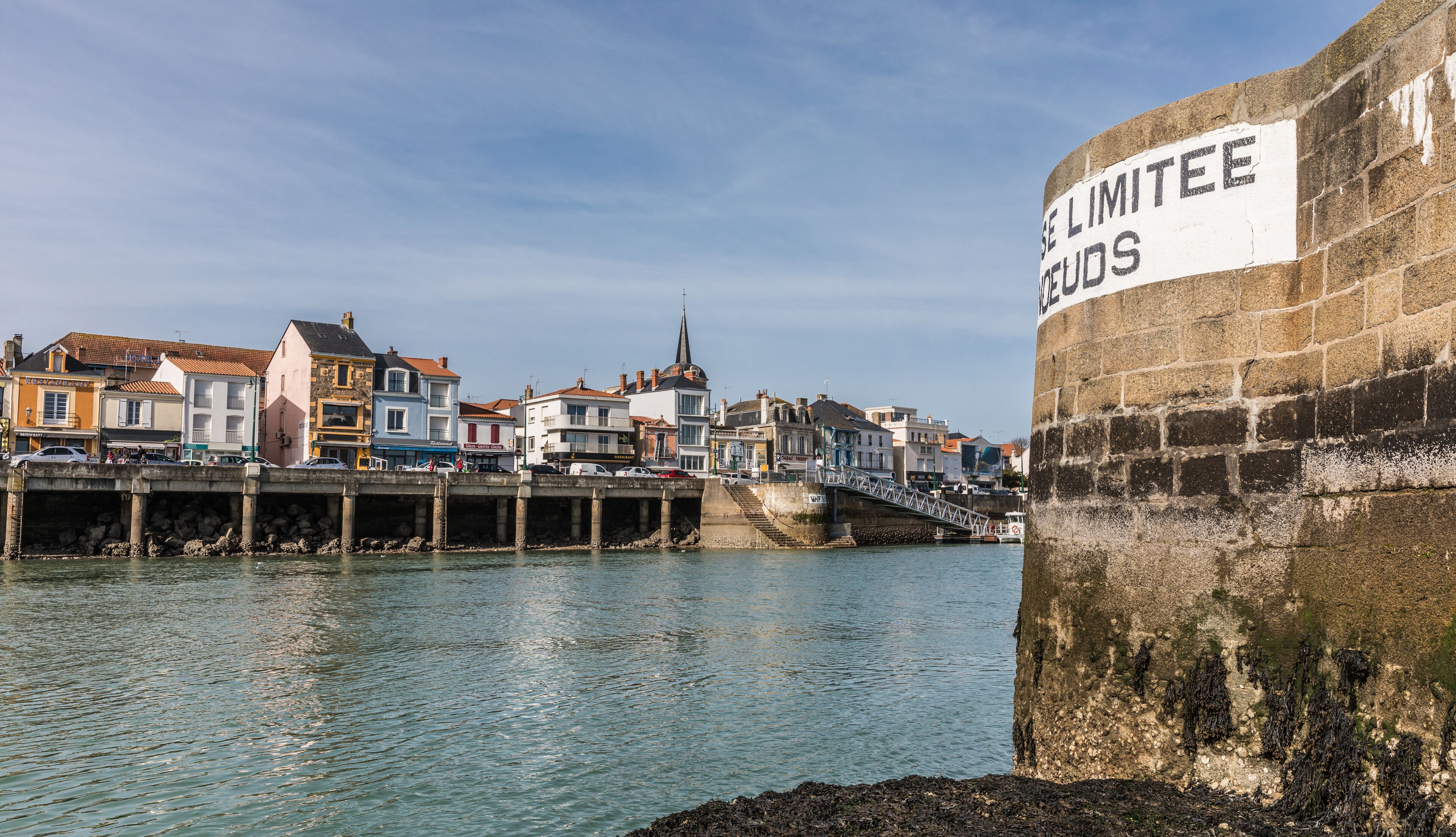 A harbor with a stone wall labeled "LIMITTEE GOODS" and a bridge leading to a town with colorful buildings.