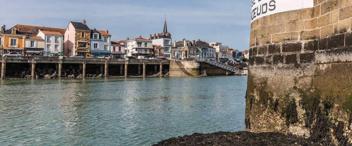 A harbor with a stone wall labeled "LIMITTEE GOODS" and a bridge leading to a town with colorful buildings.