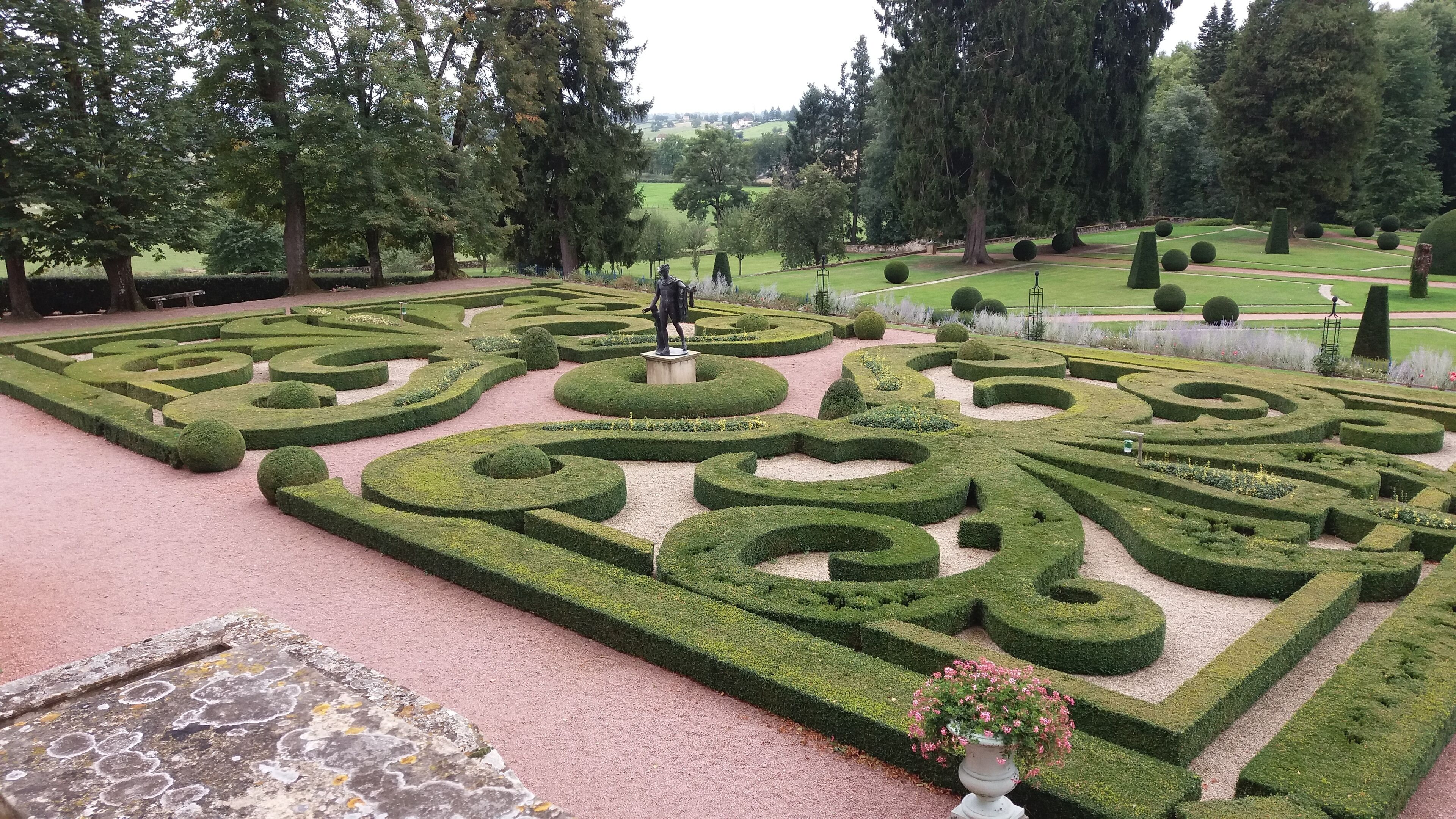 A formal garden with geometric hedge patterns, a statue, and a variety of trees.