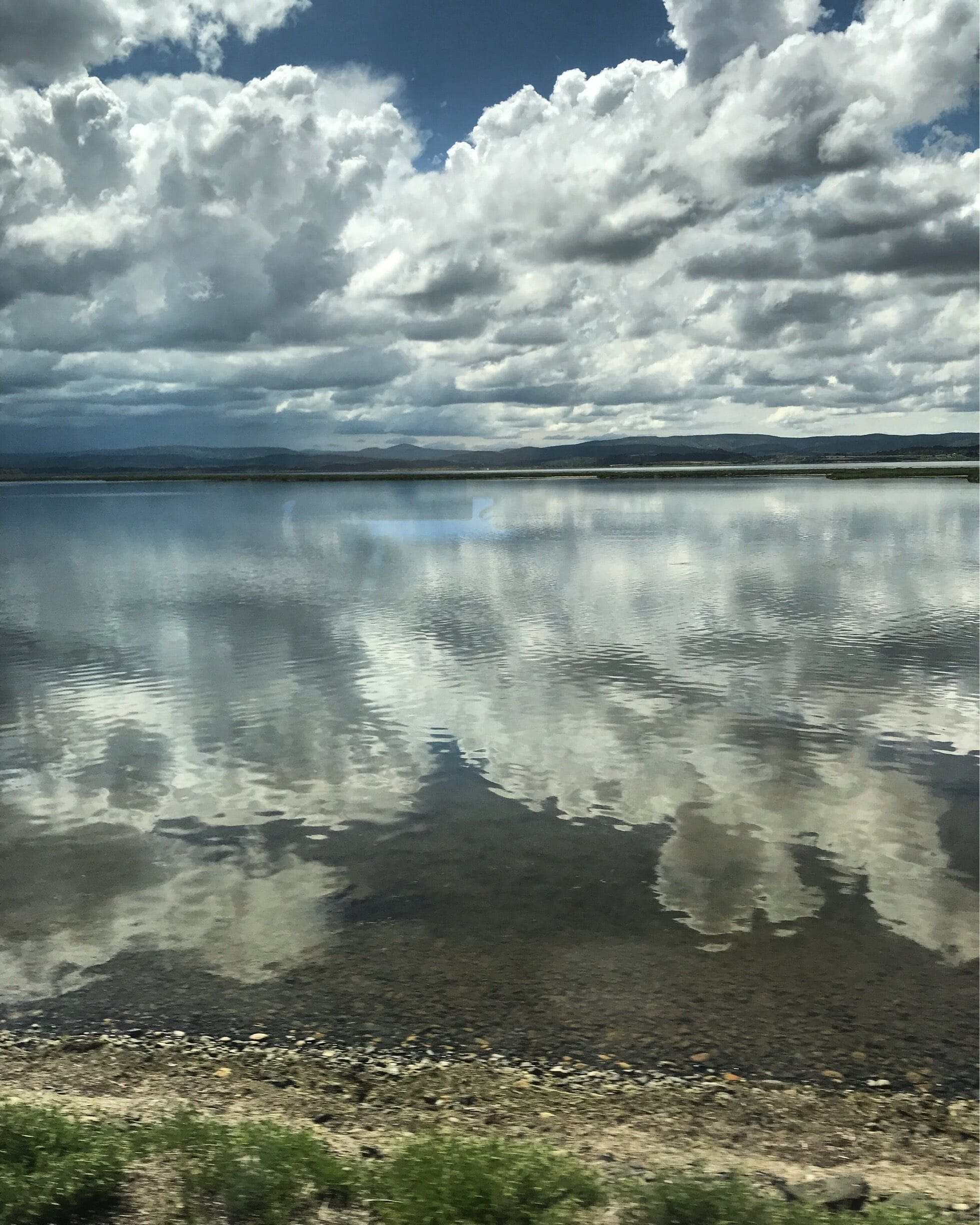 A calm lake with clear reflections of clouds, surrounded by a rocky shoreline and distant mountains.