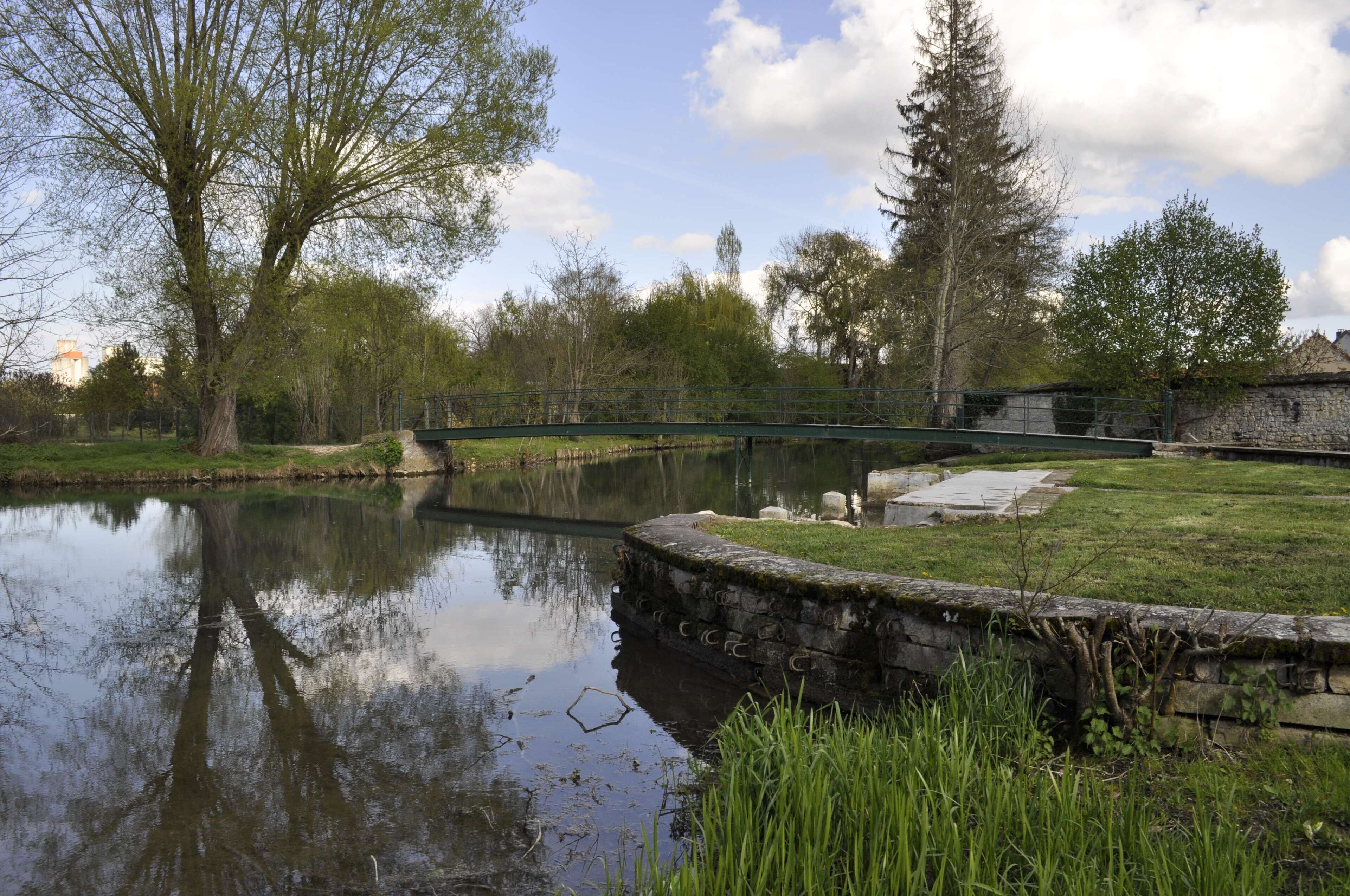 A calm pond with a stone bridge, surrounded by trees and a stone wall.