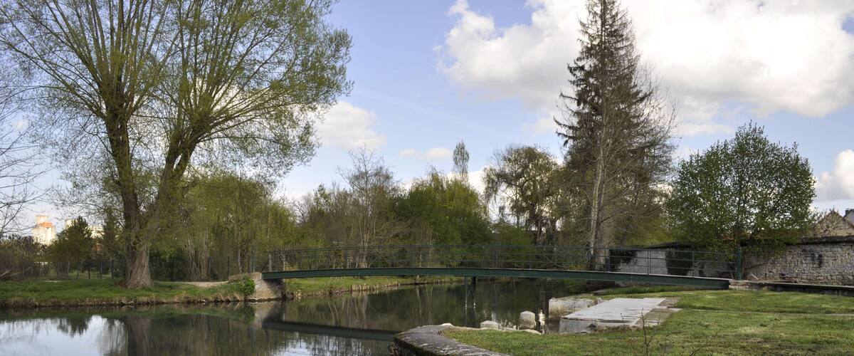 A calm pond with a stone bridge, surrounded by trees and a stone wall.
