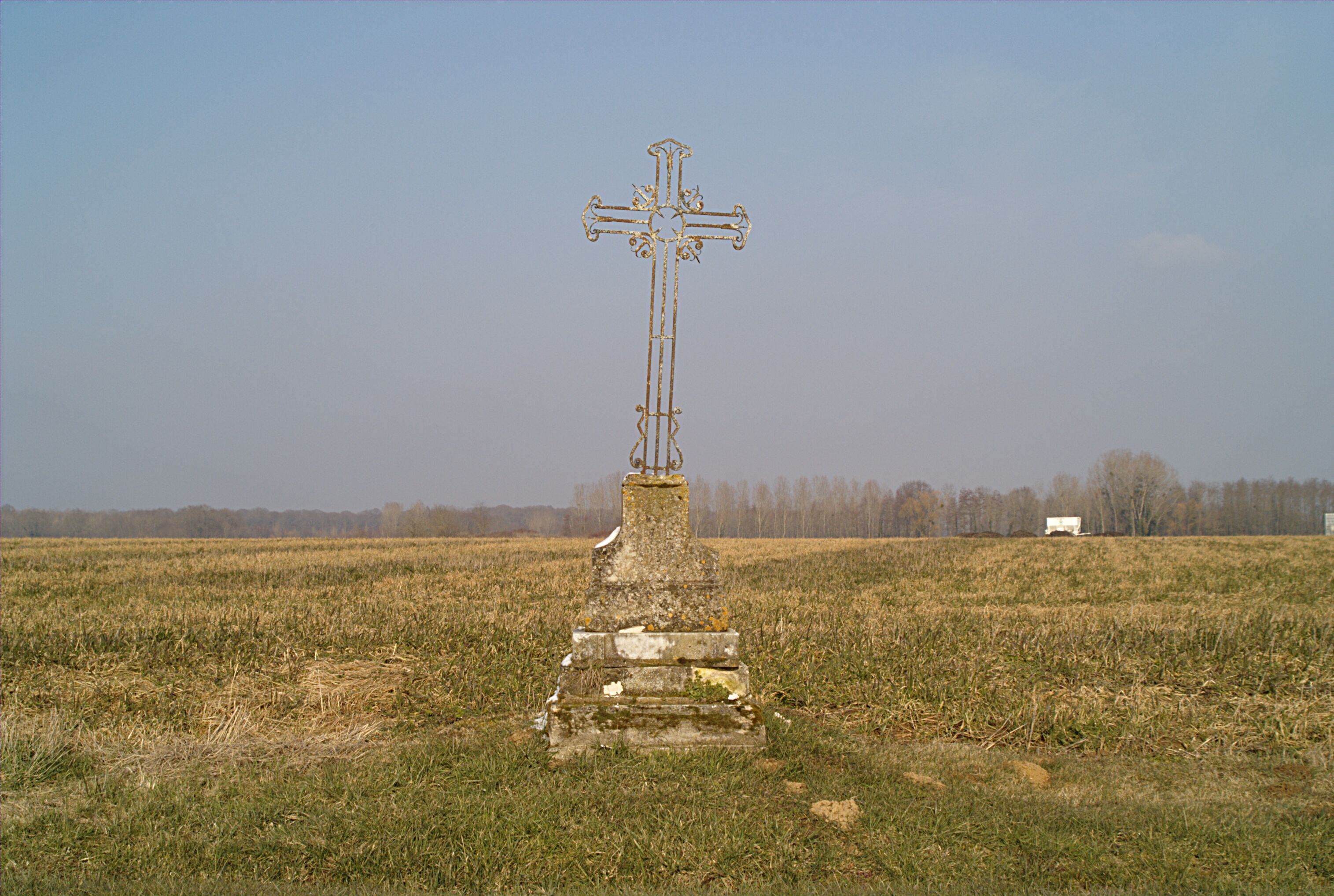 A cross monument in a field with a distant truck and leafless trees.