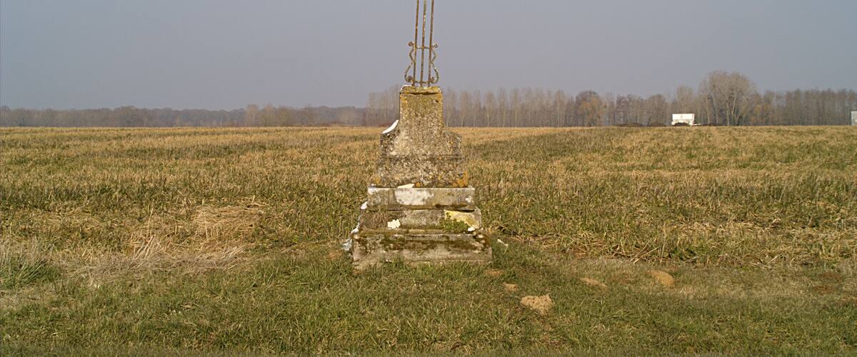 A cross monument in a field with a distant truck and leafless trees.