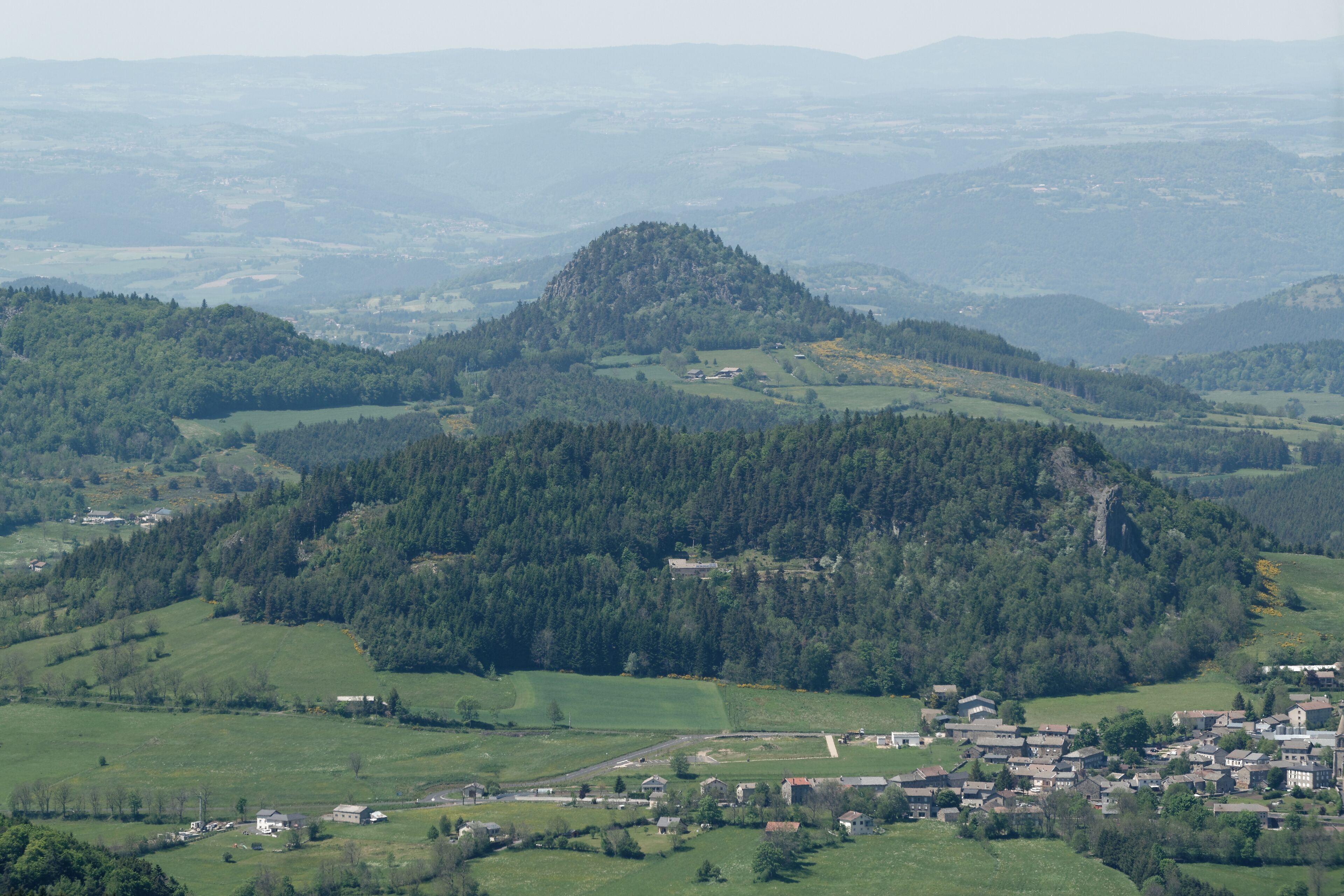 A hilly landscape with a village nestled in a valley, surrounded by dense forests and rolling green fields.