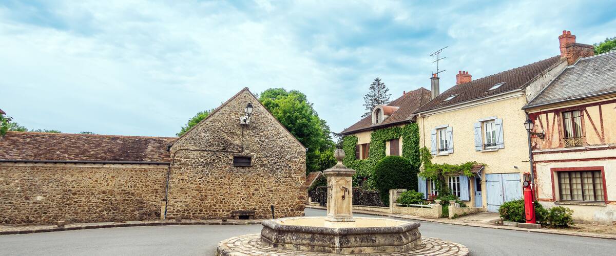 A stone building with a bell tower, a fountain, and traditional houses with shutters.