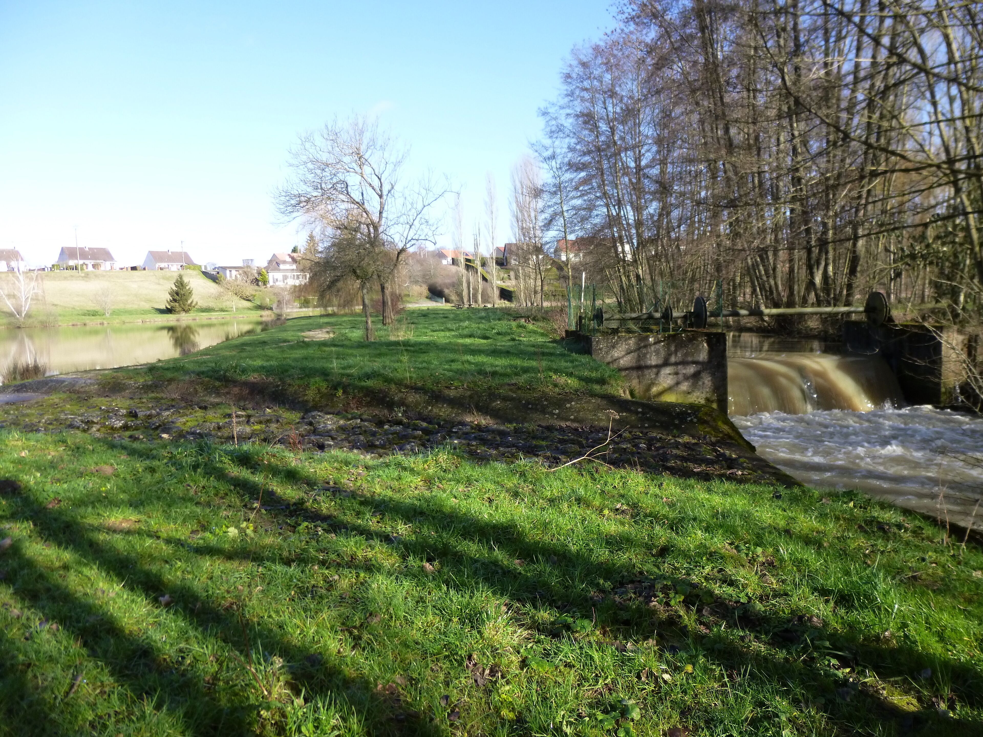La Chapelle-sur-Aveyron, Loiret, Centre, France. Communal pond, south-east side. The Aveyron river flows on the right here. The pond's overflow runs over the path which goes around the pond, to join with the Aveyron river just after a weir.