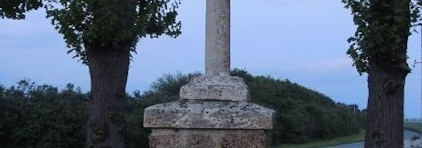 Croix BoissĂ©e, haute de 20 ou 30 cm et cerclĂ©e, en haut d'un cylindre d'1m50 environs posĂ© sur un piĂ©destale parallĂ©lĂ©pipĂ©dique de 75/80 cm environs posĂ© au centre du second disque, le plus petit, lui mĂȘme centrĂ© et au dessus du plus grand disque de pierre entre deux arbres Ă l'arriĂšre plan et la route derriĂšre encore