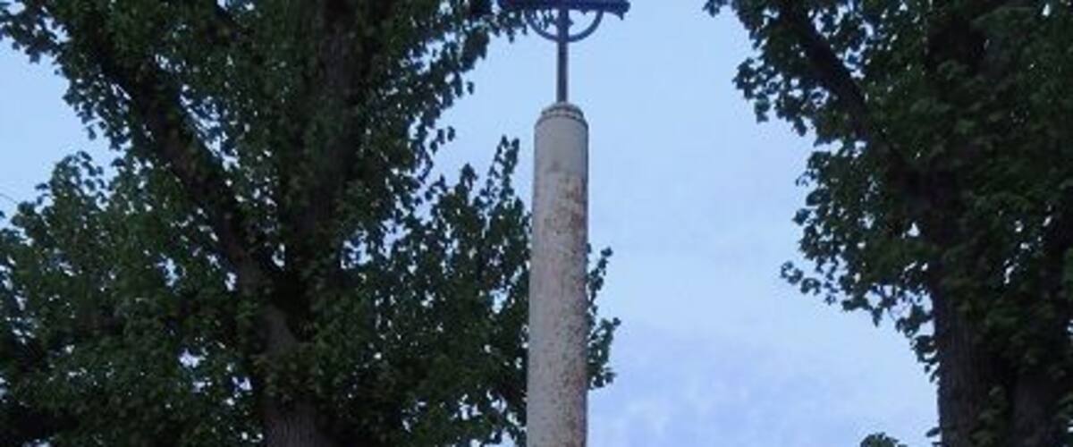 A stone monument with a cross on top, surrounded by a circular stone base, set in a grassy area with trees.