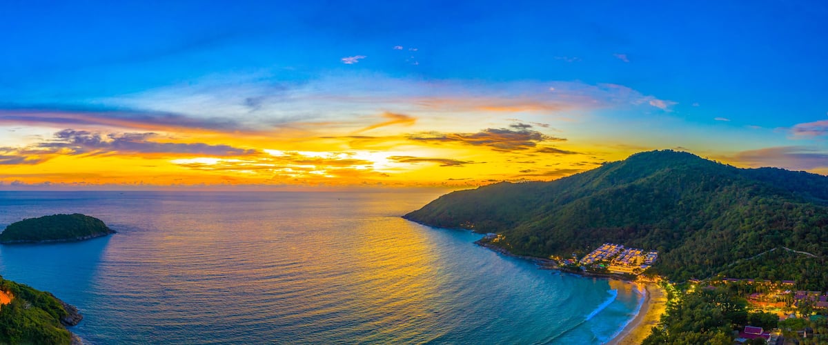 A coastal landscape with a sunset, a small island, and a beach with buildings.