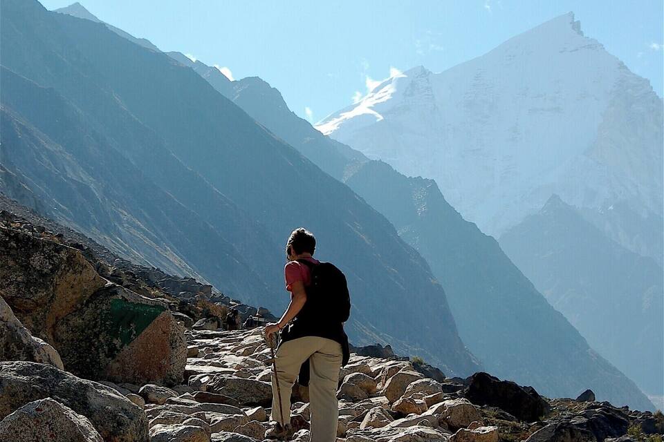 A hiker on a rocky mountain path with snow-capped peaks in the background.