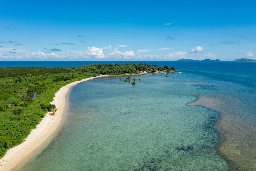 Un paysage côtier avec une plage de sable, des eaux turquoise et limpides, et une île boisée à l’horizon.