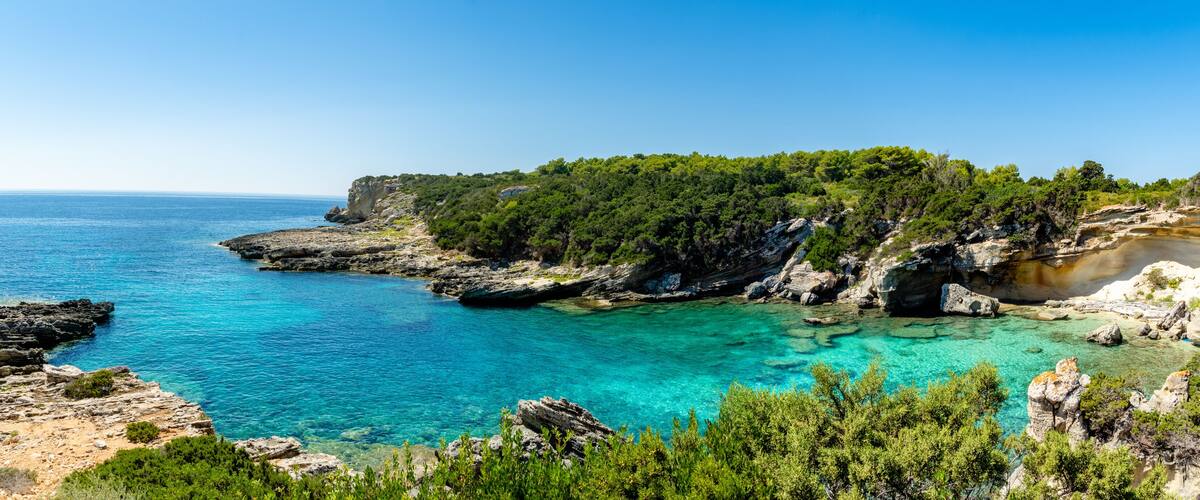 A coastal landscape with clear turquoise waters, rocky cliffs, and dense green vegetation.