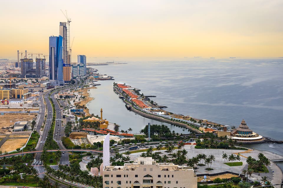 A coastal cityscape with a prominent skyscraper, a mosque, and a waterfront promenade.
