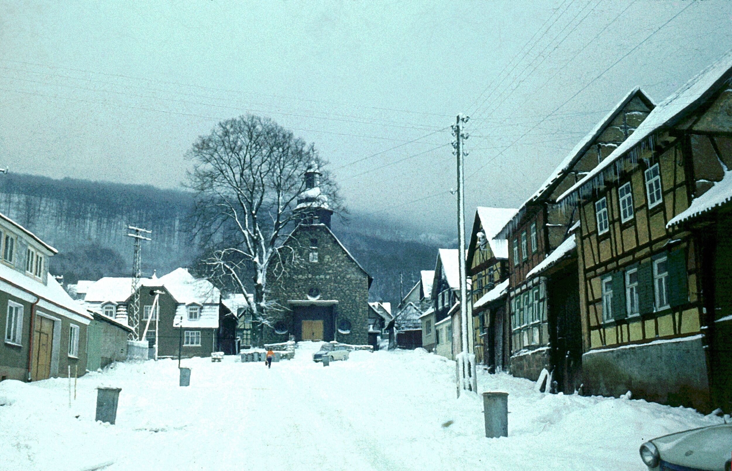 A snow-covered village street with traditional half-timbered houses, a stone church, and a large tree in the background.