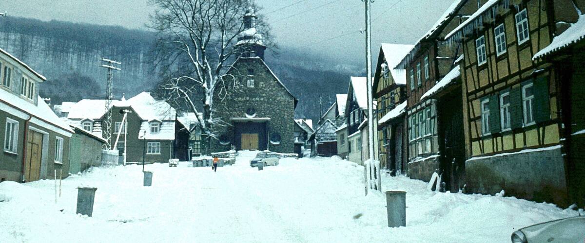 A snow-covered village street with traditional half-timbered houses, a stone church, and a large tree in the background.