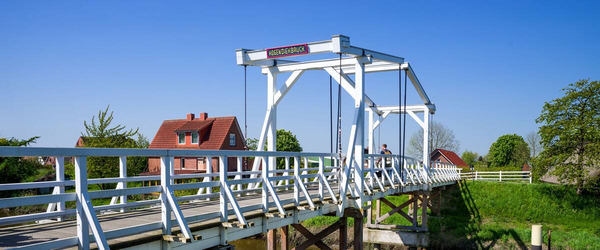 A white metal bridge over a calm river with a red-roofed house and a white fence in the background.