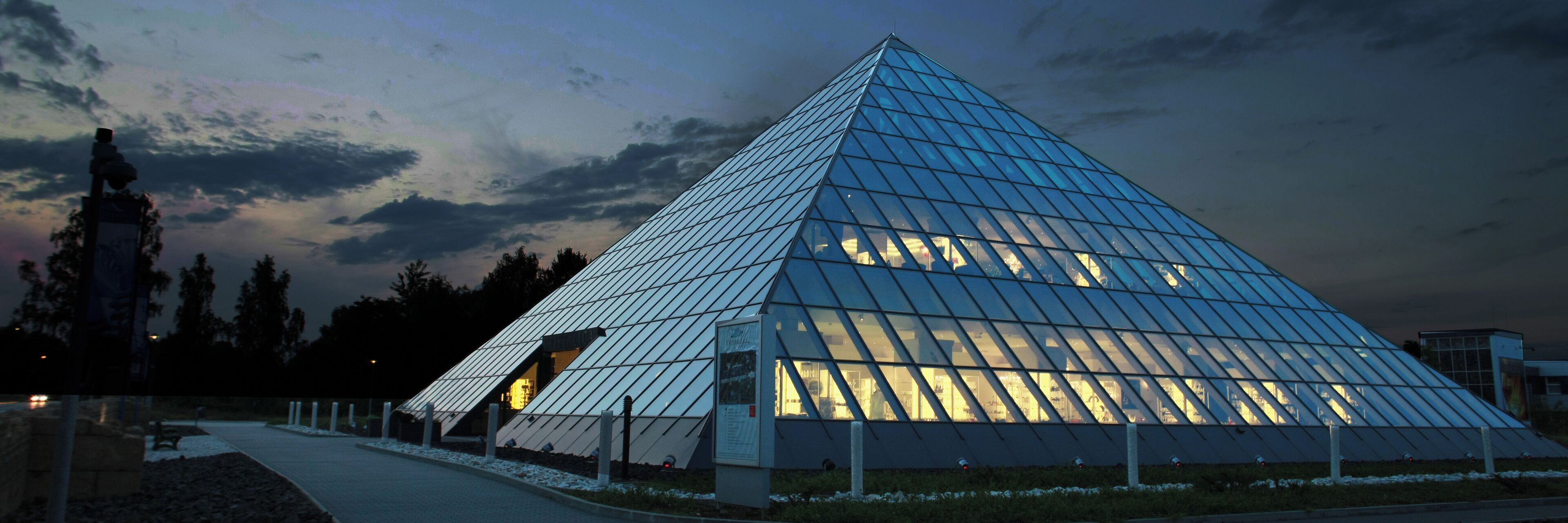 A large glass pyramid structure with a sloped roof, surrounded by a pathway and trees.