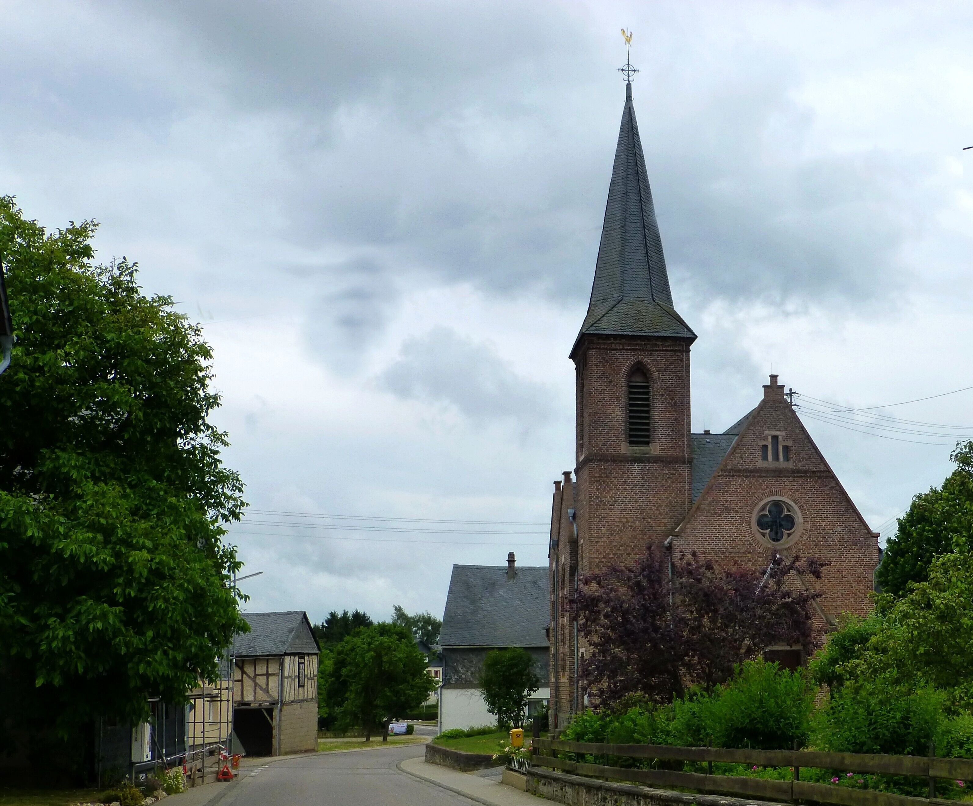 A church with a tall steeple and a weather vane, surrounded by trees and a small building.