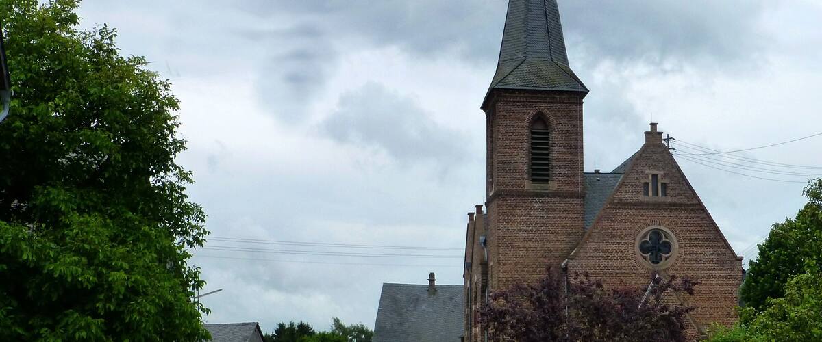 A church with a tall steeple and a weather vane, surrounded by trees and a small building.
