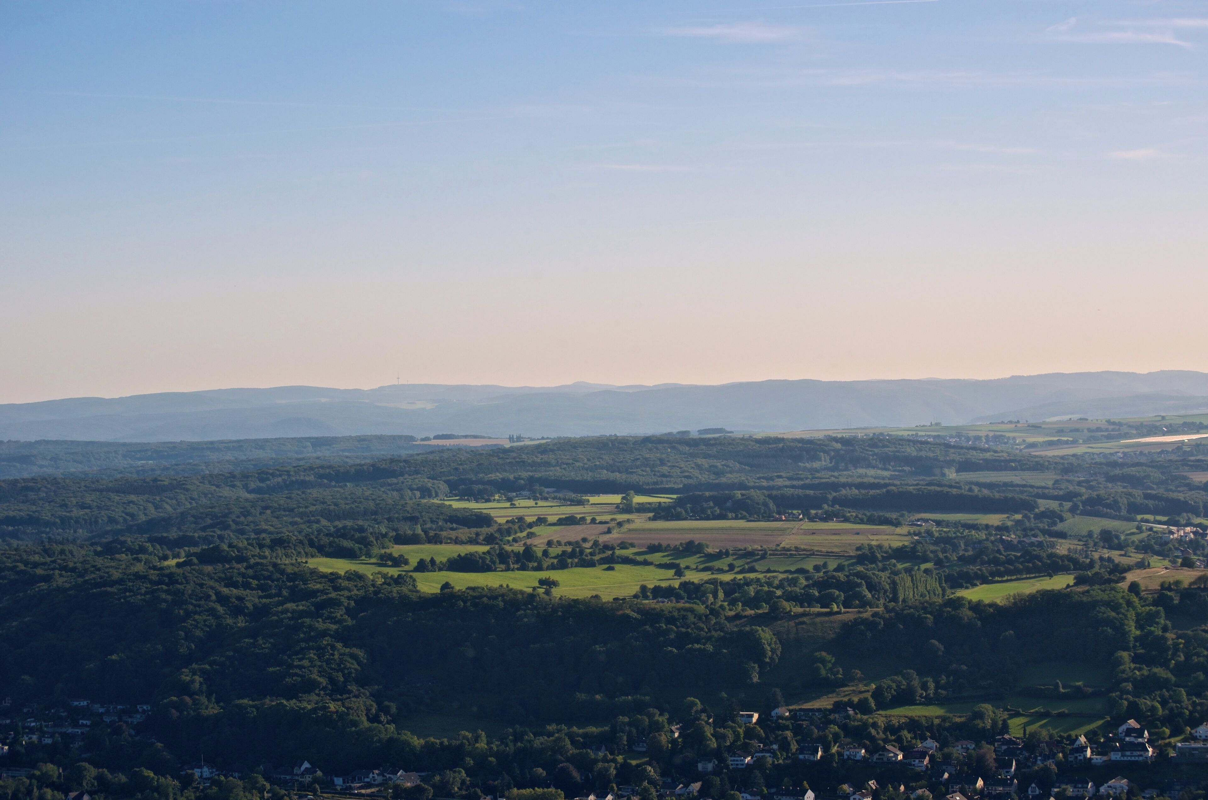 A panoramic view of a lush valley with rolling hills, scattered houses, and distant mountains under a clear sky.