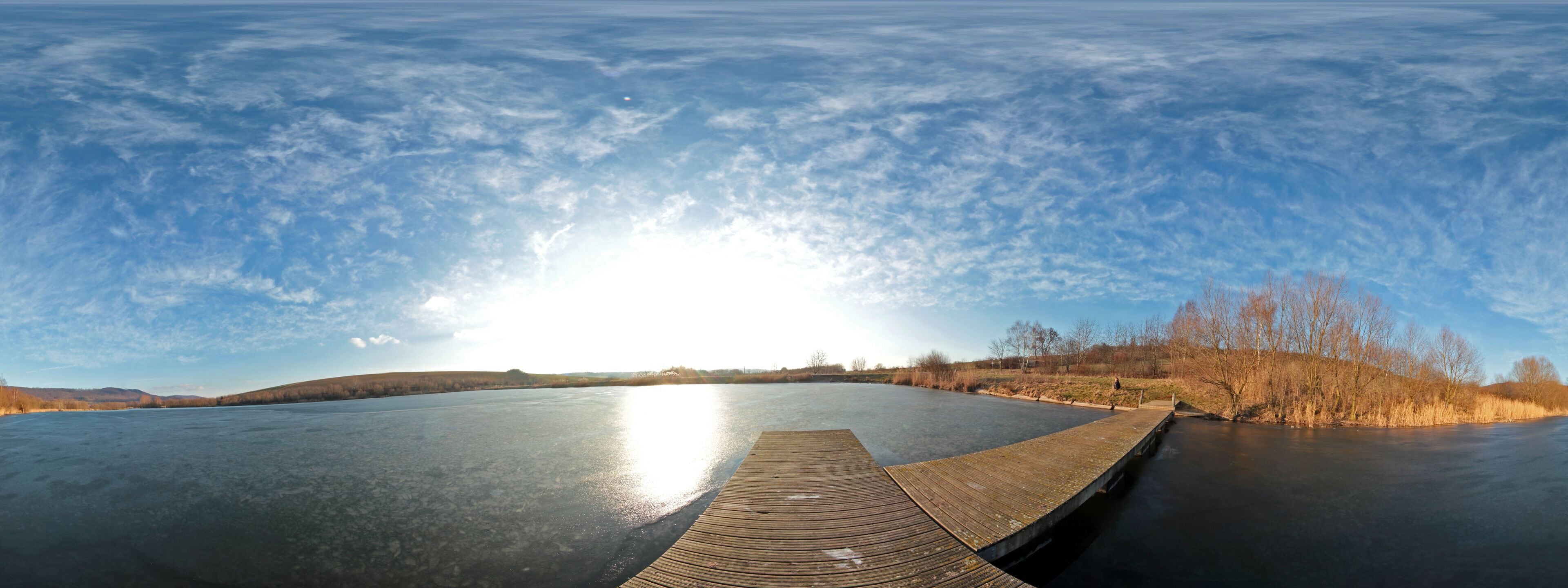 A frozen lake with a wooden dock extending into the water, surrounded by trees and hills under a clear sky.