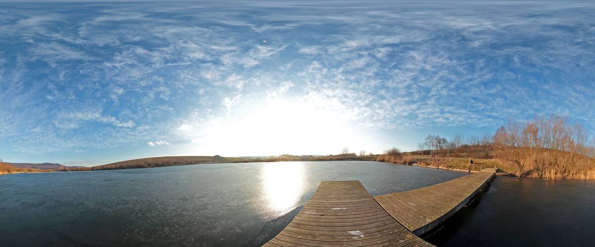 A frozen lake with a wooden dock extending into the water, surrounded by trees and hills under a clear sky.