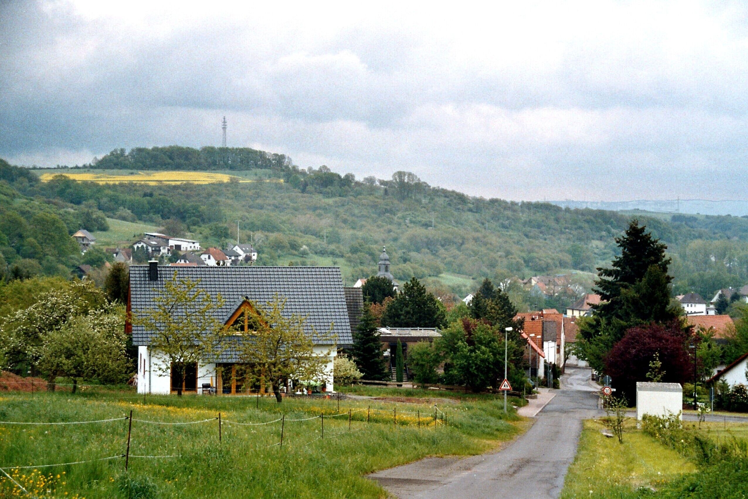 A rural road flanked by houses, a church, and a hill with a power line tower in the distance.