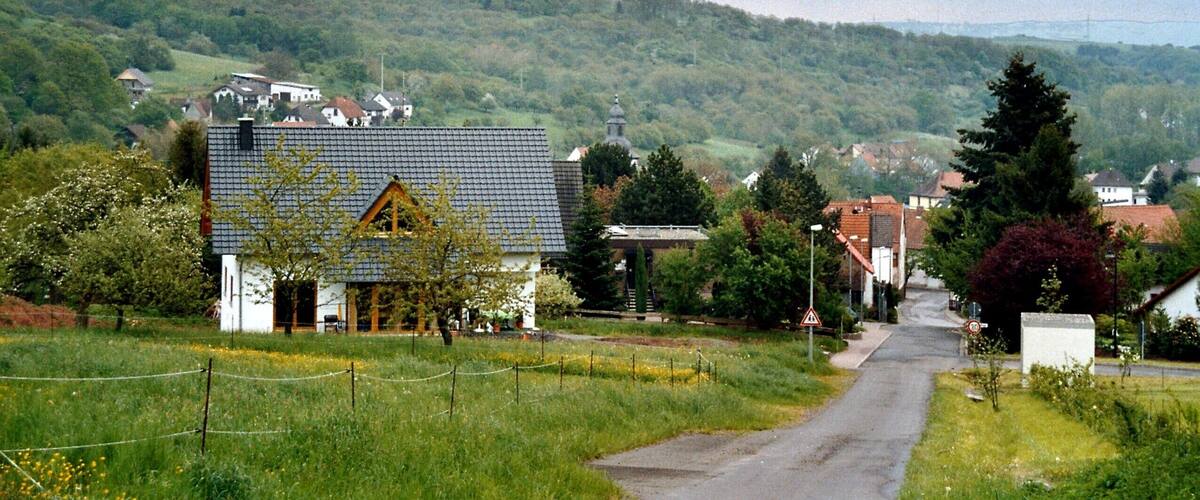 A rural road flanked by houses, a church, and a hill with a power line tower in the distance.
