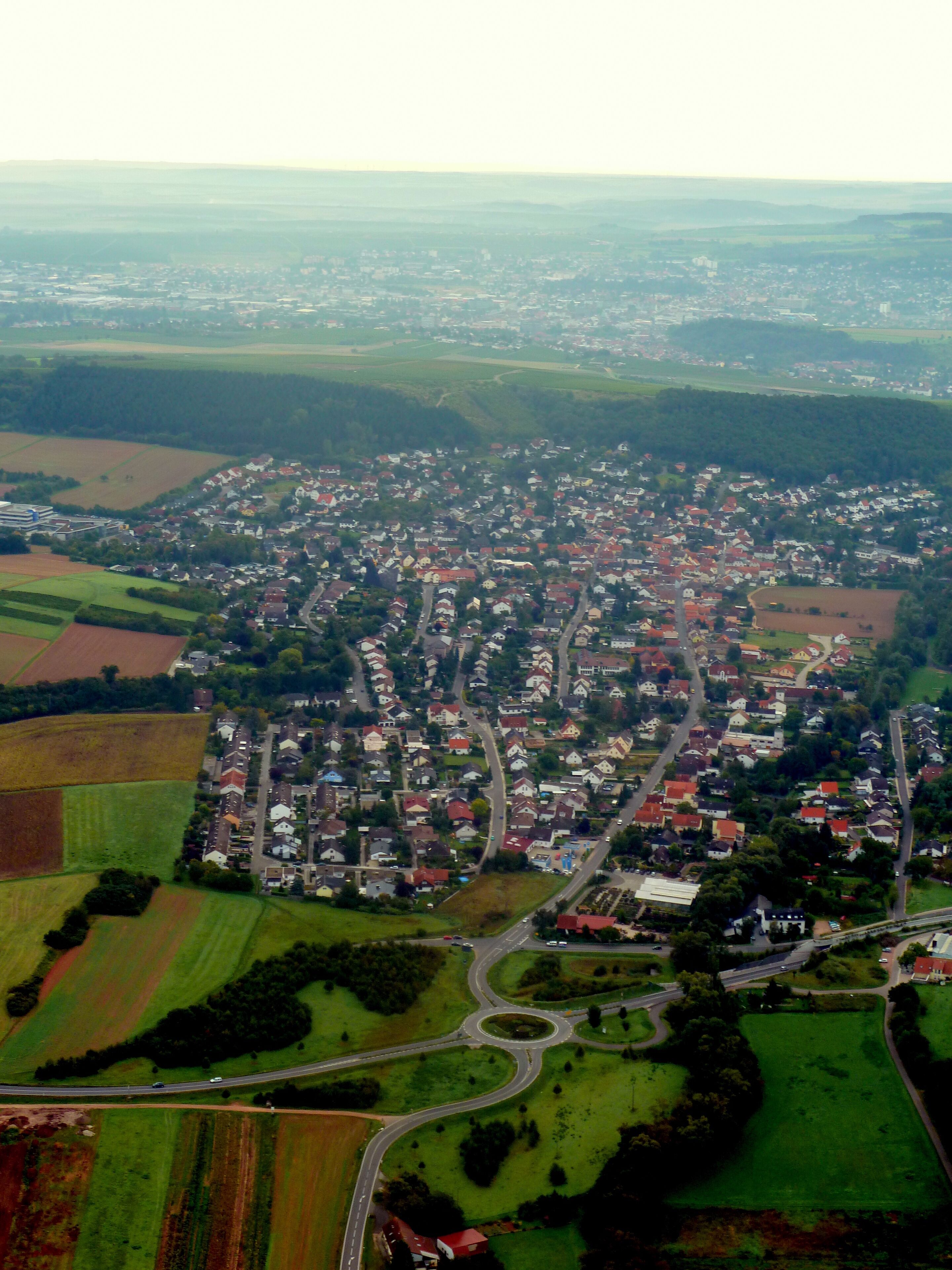 An aerial view of a town with a dense cluster of houses, surrounded by green fields and a winding road.