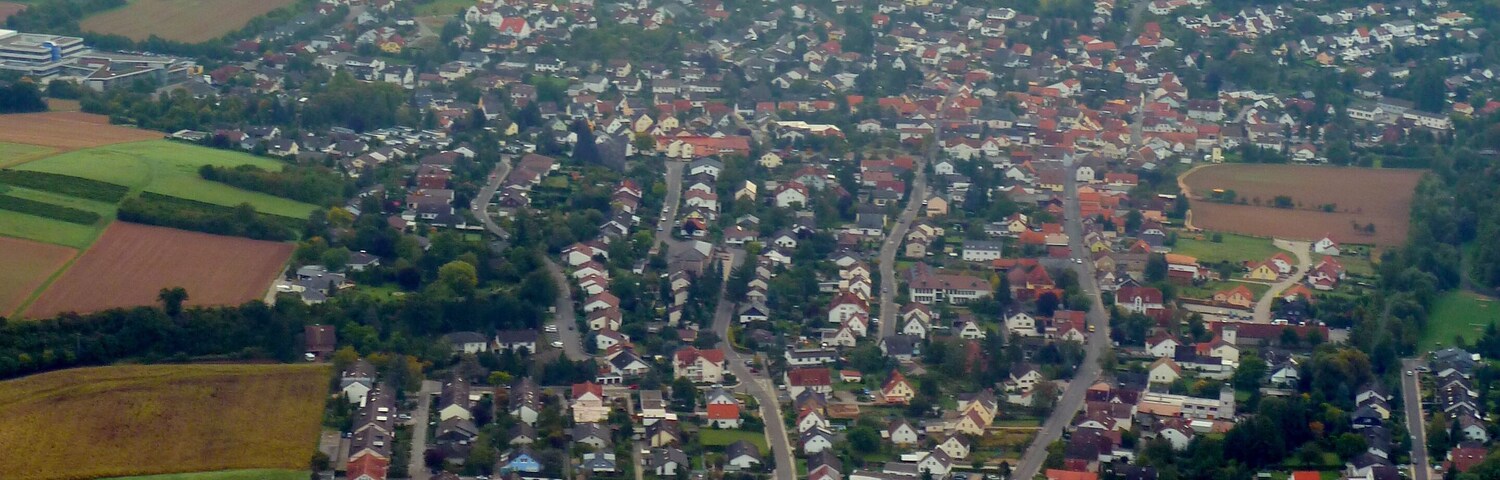 An aerial view of a town with a dense cluster of houses, surrounded by green fields and a winding road.