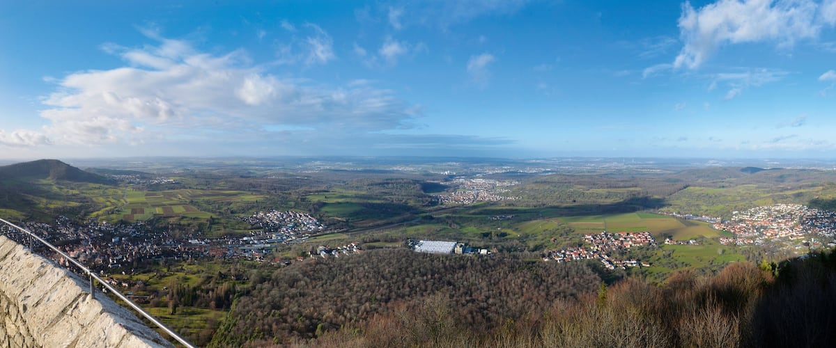 A panoramic view of a valley with a town, rolling hills, and a distant mountain range under a clear sky.